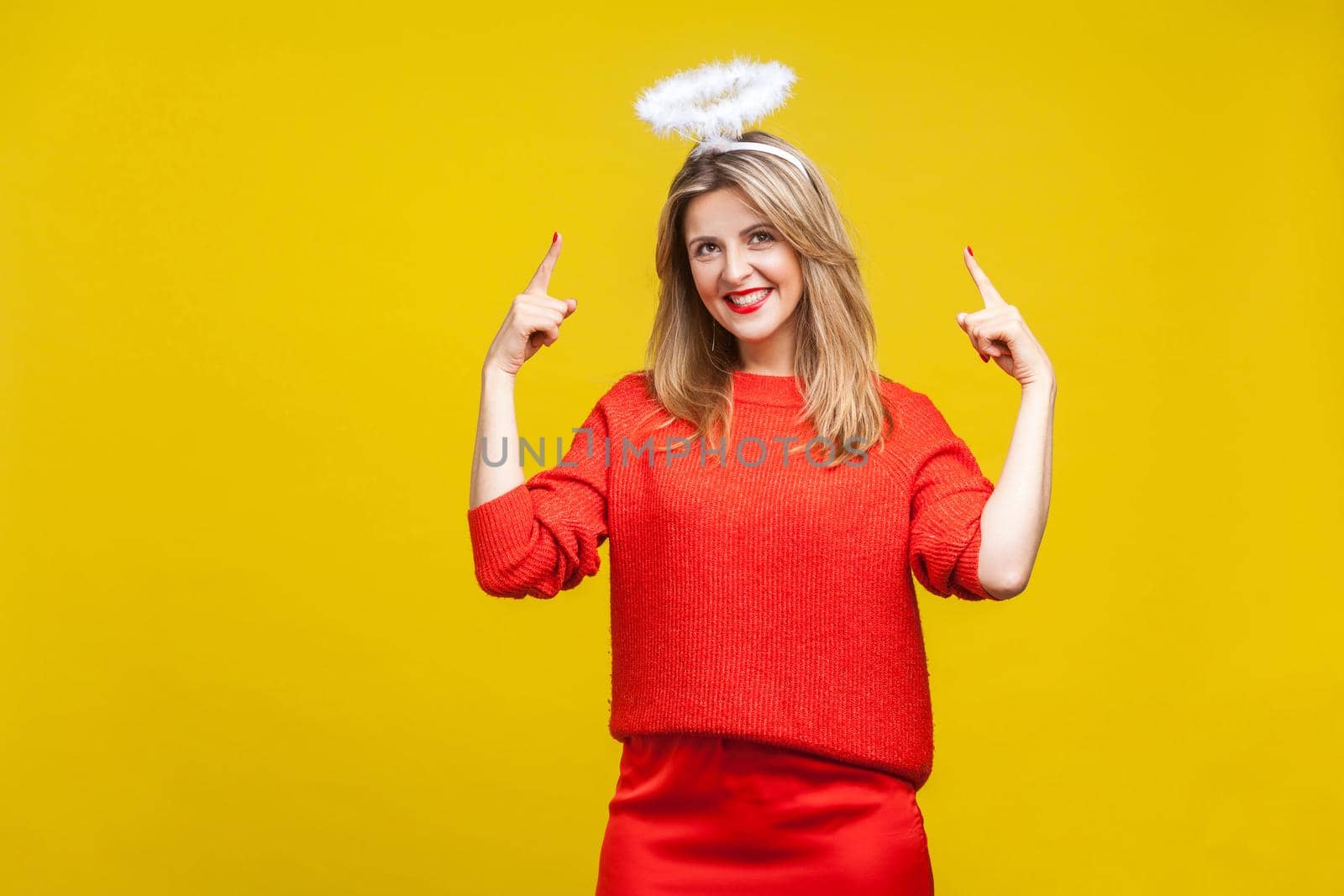 Portrait of proud adorable young woman with bright makeup in red casual clothes standing, pointing at hola on her head and smiling. indoor studio shot isolated on yellow background