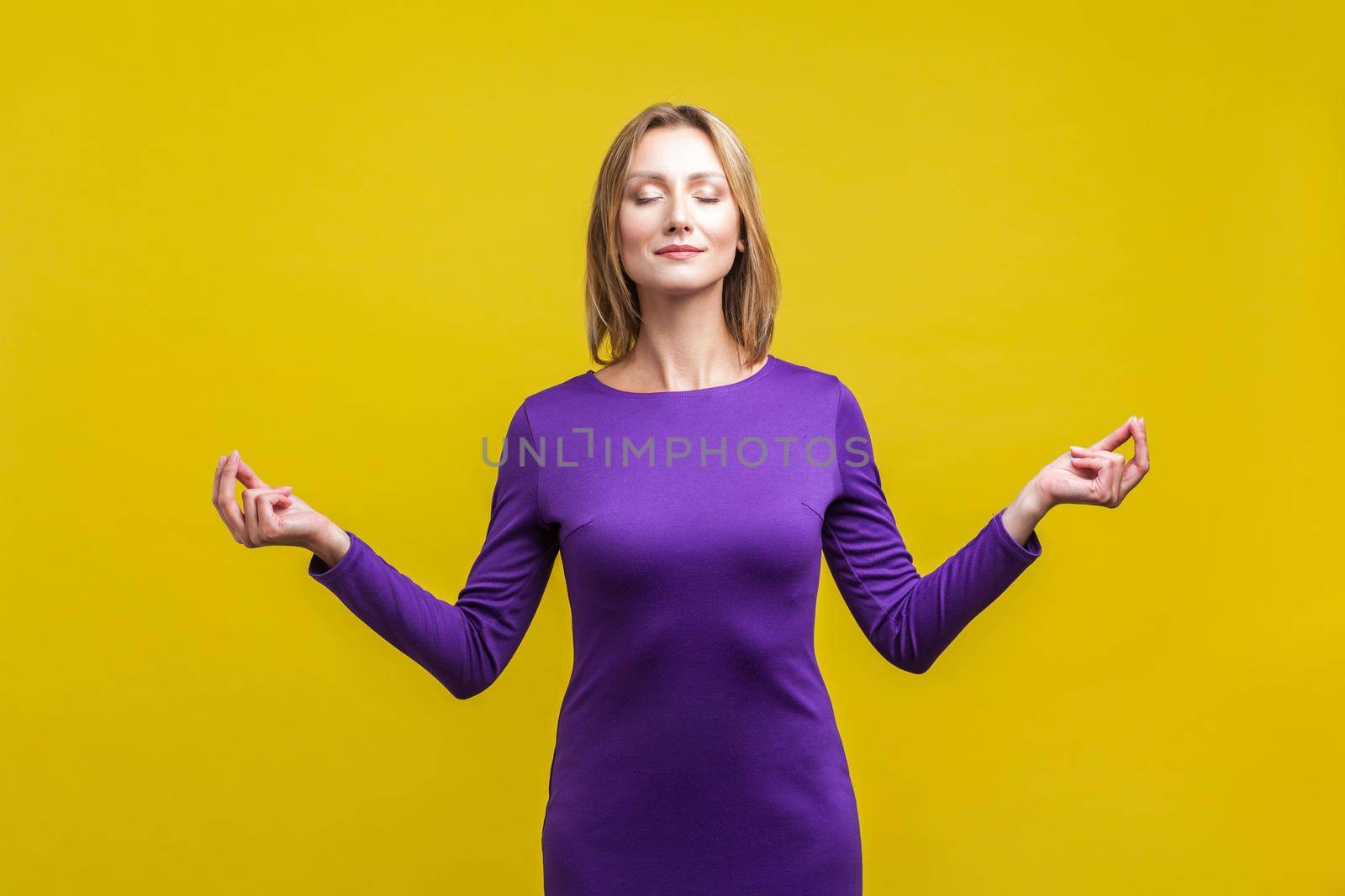 Yoga practice, harmony. Portrait of peaceful woman in purple dress standing with closed eyes and calm face meditating, holding fingers in mudra gesture. studio shot isolated on yellow background