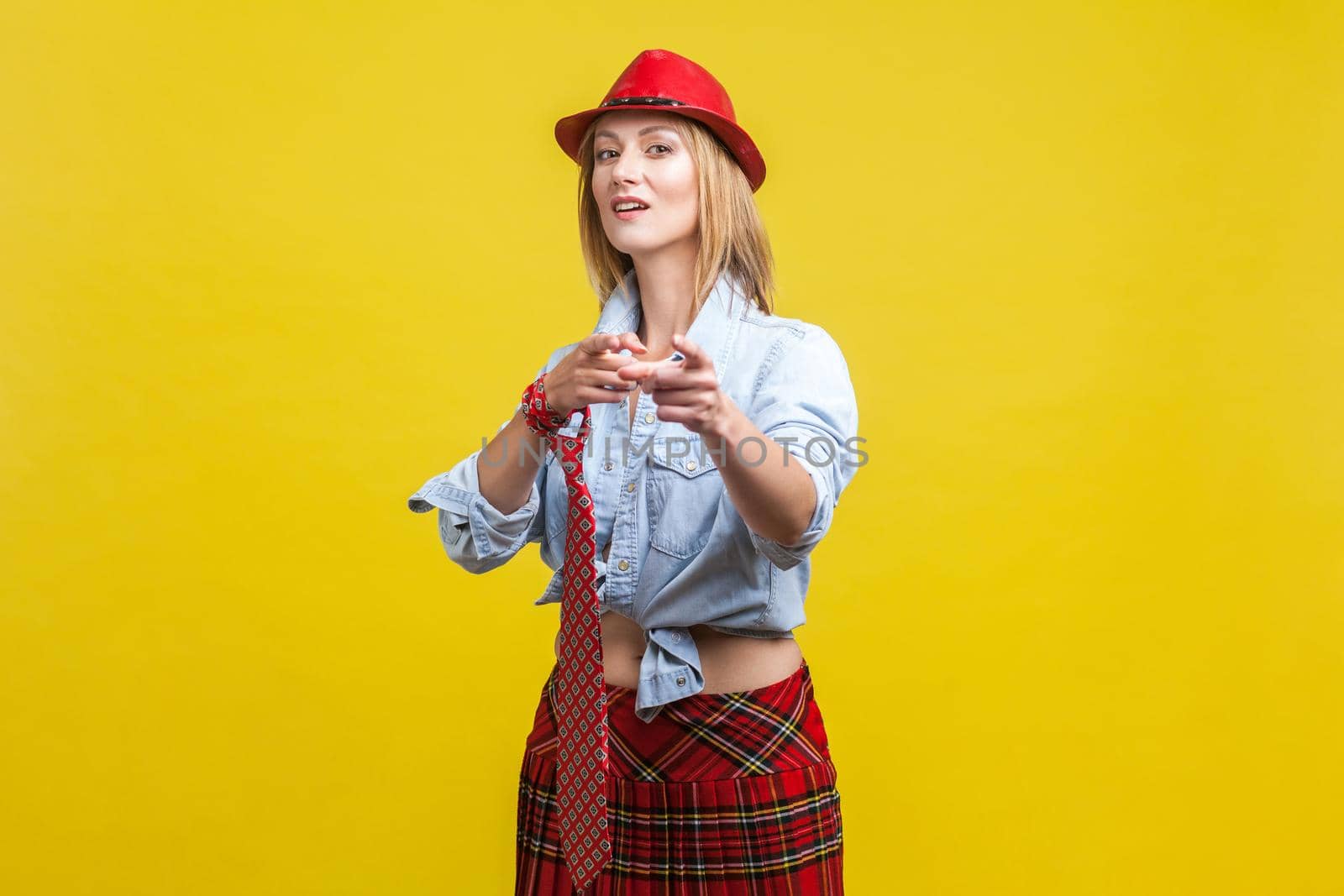 Portrait of beautiful glamorous woman wearing stylish red hat, school skirt and tie knotted on hand, pointing fingers at camera. showing hey you gesture. studio shot isolated on yellow background