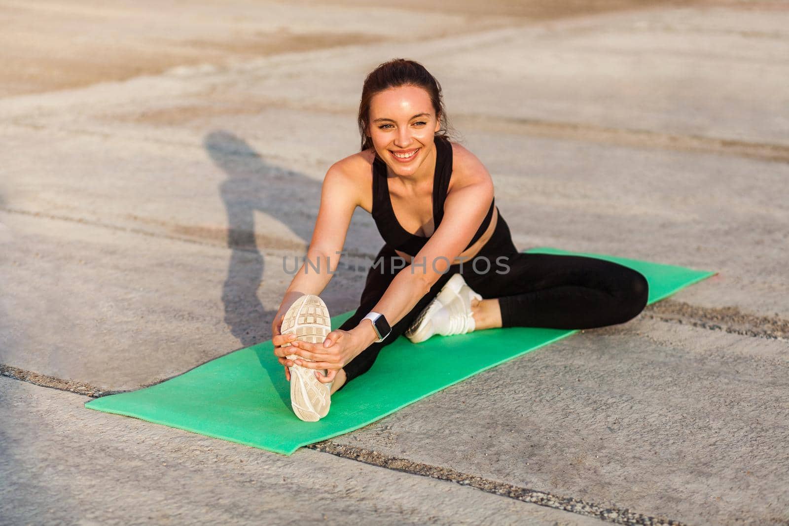 Cheerful smiling athletic woman in tight sportswear, black pants and top, practicing yoga, doing Head-to-Knee Pose, touching toes, stretching leg and back muscles. Health care, sport activity outdoor