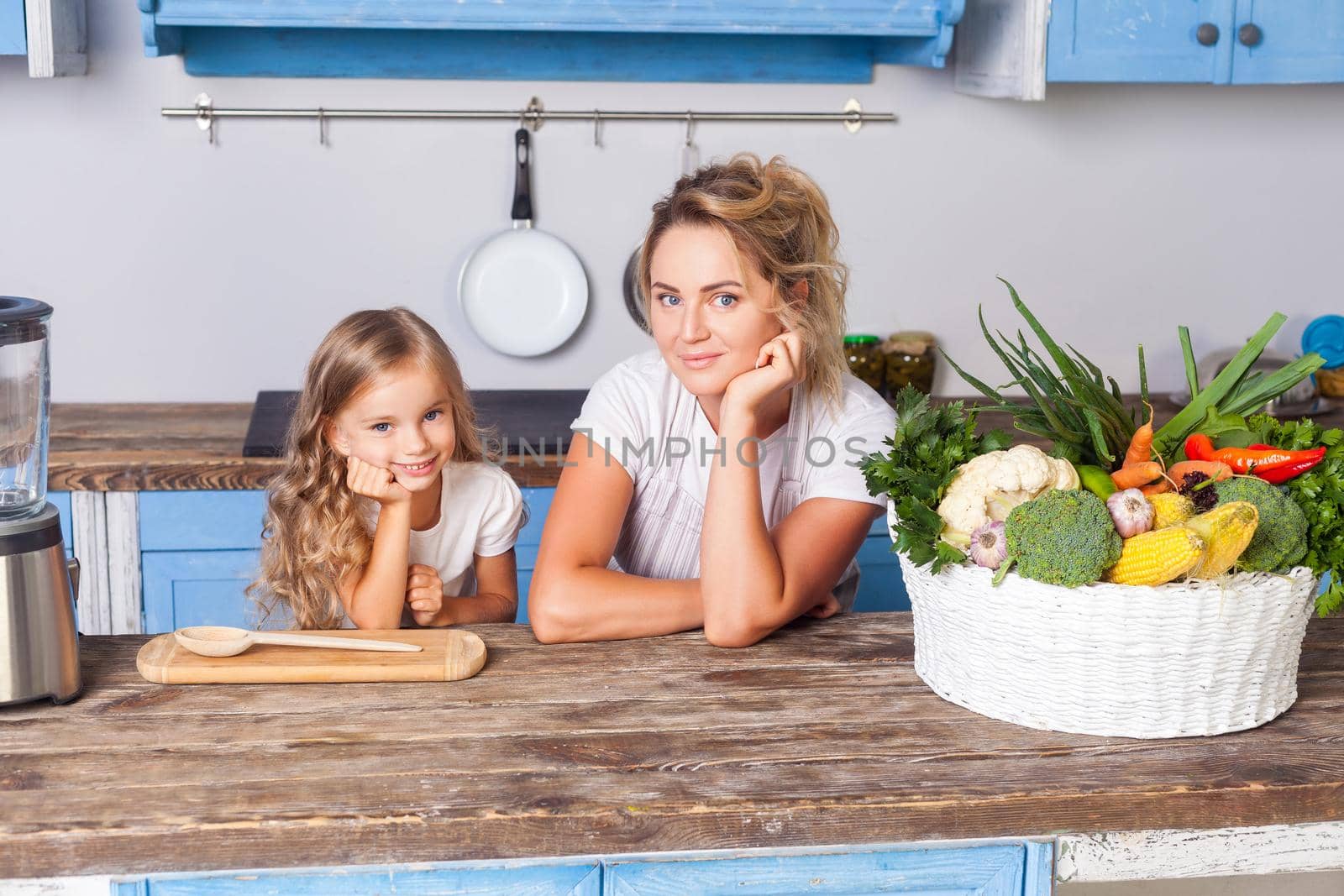 Young cheerful mother and little daughter leaning on table, standing in kitchen with modern furniture, basket of fresh vegetables on table, eco-friendly vegetarian food, vegan nutrition, healthcare