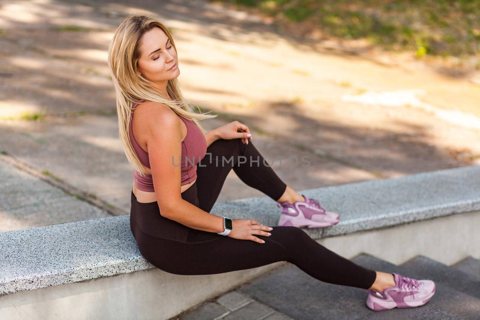 Sexy sportswoman sitting on stairs with her eyes closed and pleased expression, taking break relaxing, feeling fit attractive after trainings outdoor. Concept of beauty and health with sport lifestyle