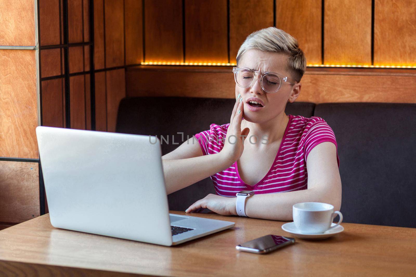 Portrait of unwell young adult woman with short blonde hair in pink t-shirt and eyeglasses sitting in cafe and touching cheek with hand have a high pain, toothache and dentistry. Indoor, health care