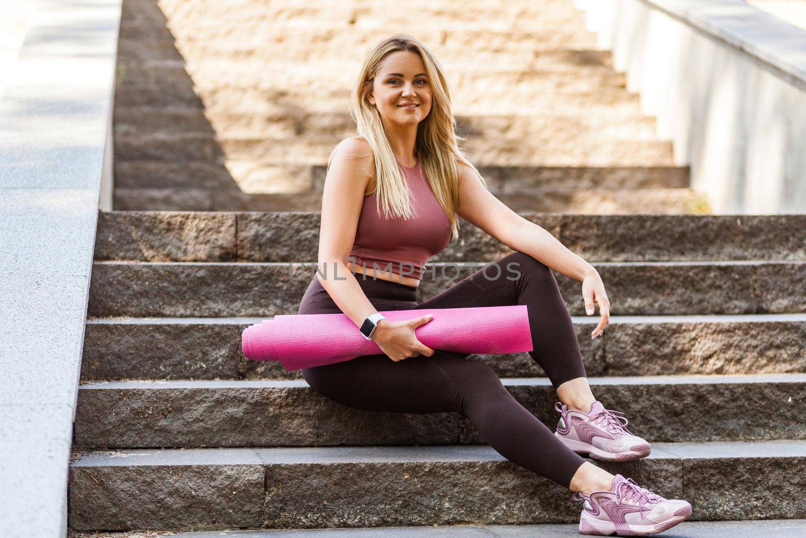 Happy fit blond girl in yoga pants sitting on stairs, holding mat and smiling with pleased satisfied expression, relaxing taking break after sport training outdoor. Enjoying healthy active lifestyle