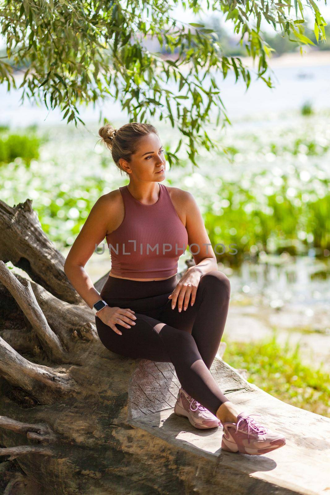 Sexy sportive woman in tight yoga pants and top sitting in forest taking rest after jogging, looking away with pensive serious concentrated expression. Relaxation after workouts outdoor summer day