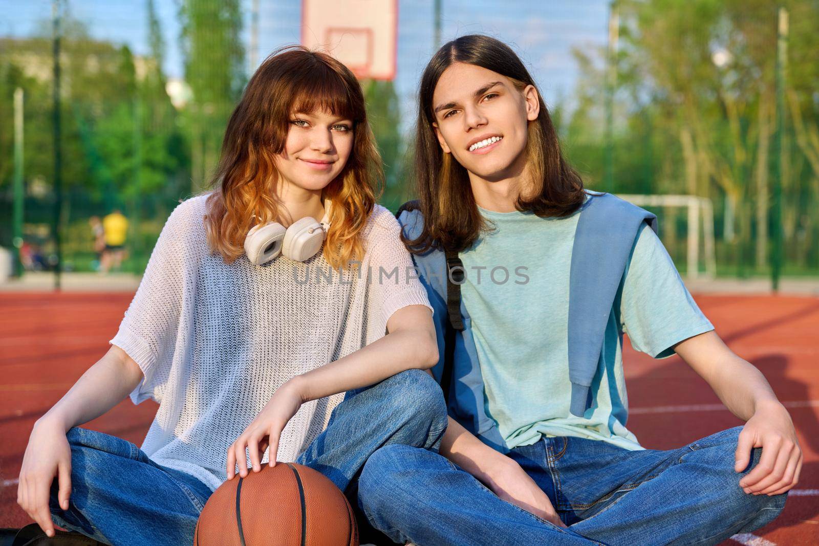 Portrait of couple friends teenagers guy and girl looking at camera, sitting on school basketball court. Teenage students with backpacks, ball in sunset. Active healthy sport lifestyle, youth concept