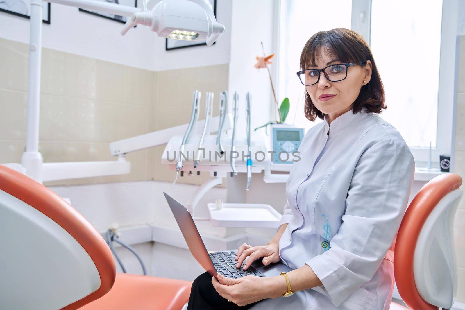 Portrait of dentist doctor sitting in office with laptop in her hands looking at camera. Mature smiling female nurse in dental clinic. Dentistry, medicine, health care, profession, stomatology concept