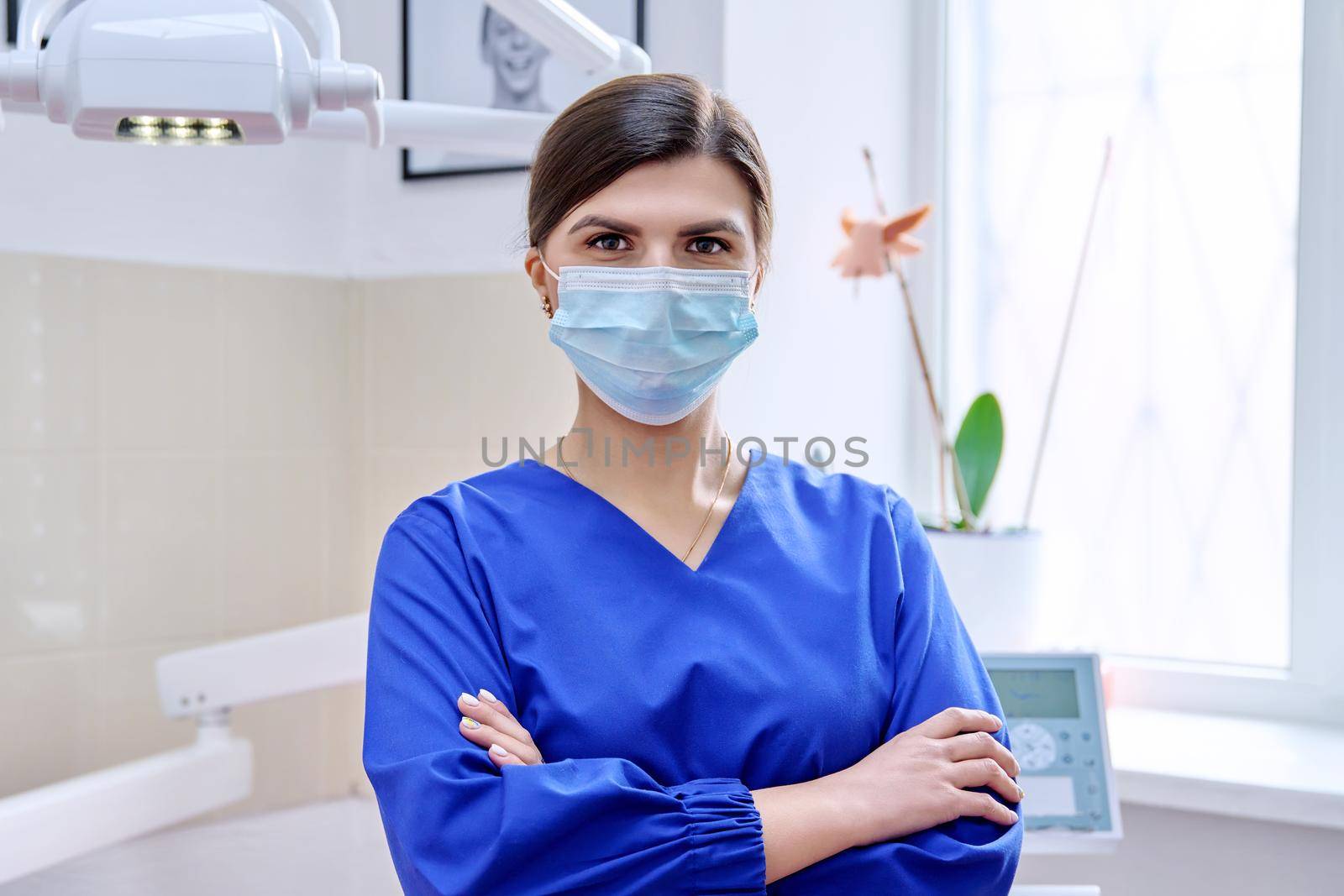 Portrait of confident female dentist doctor in office looking at camera. Young female in medical protective mask in blue uniform. Dentistry, medicine, health care, profession, stomatology