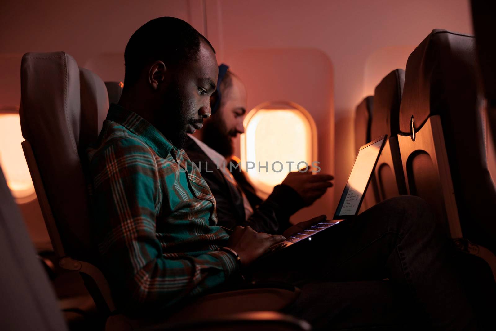 African american entrepreneur working on laptop during sunset, flying by airplane on commercial flight with international airline service. Male tourist travelling on work trip or holiday.