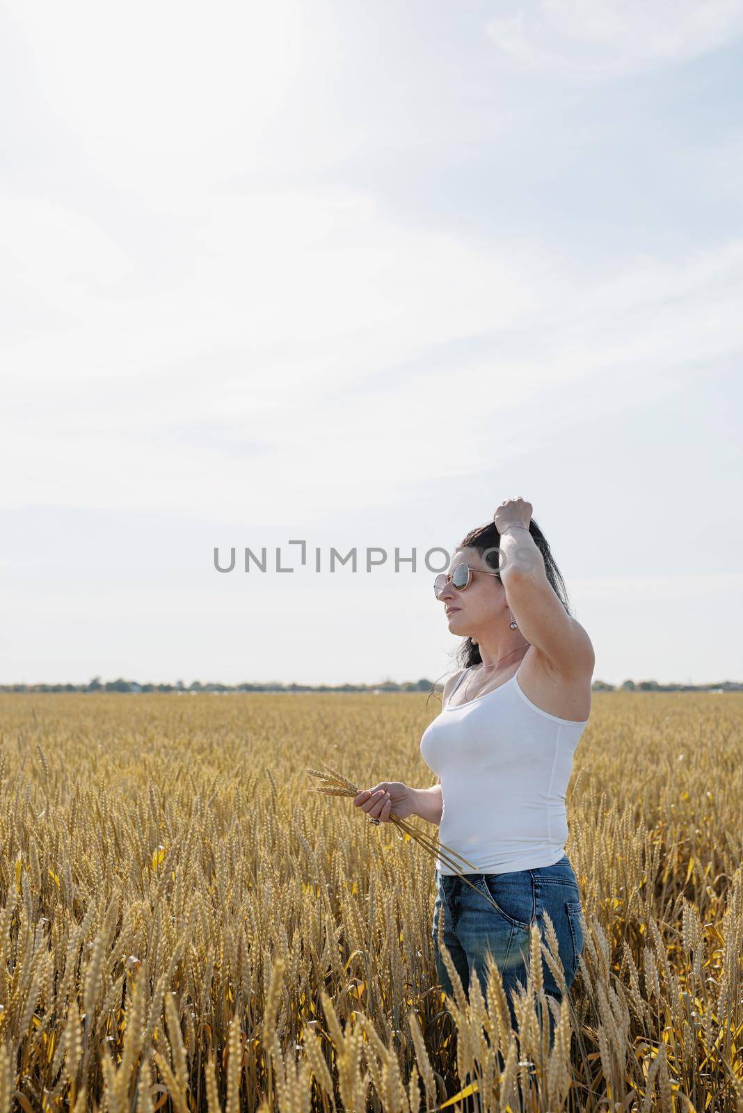 Mid adult brunette woman walking across golden field holding heap of rye lit by sunset light, copy space