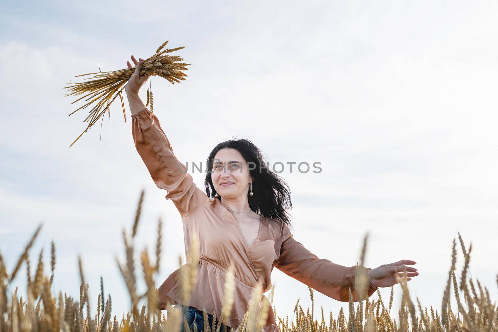 Mid adult woman in beige shirt walking across golden field holding heap of rye lit by sunset light, copy space