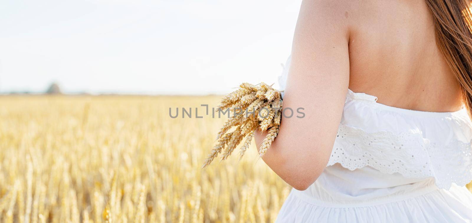 Young brunette woman walking across golden field holding heap of rye and wearing straw hat lit by sunset light, copy space