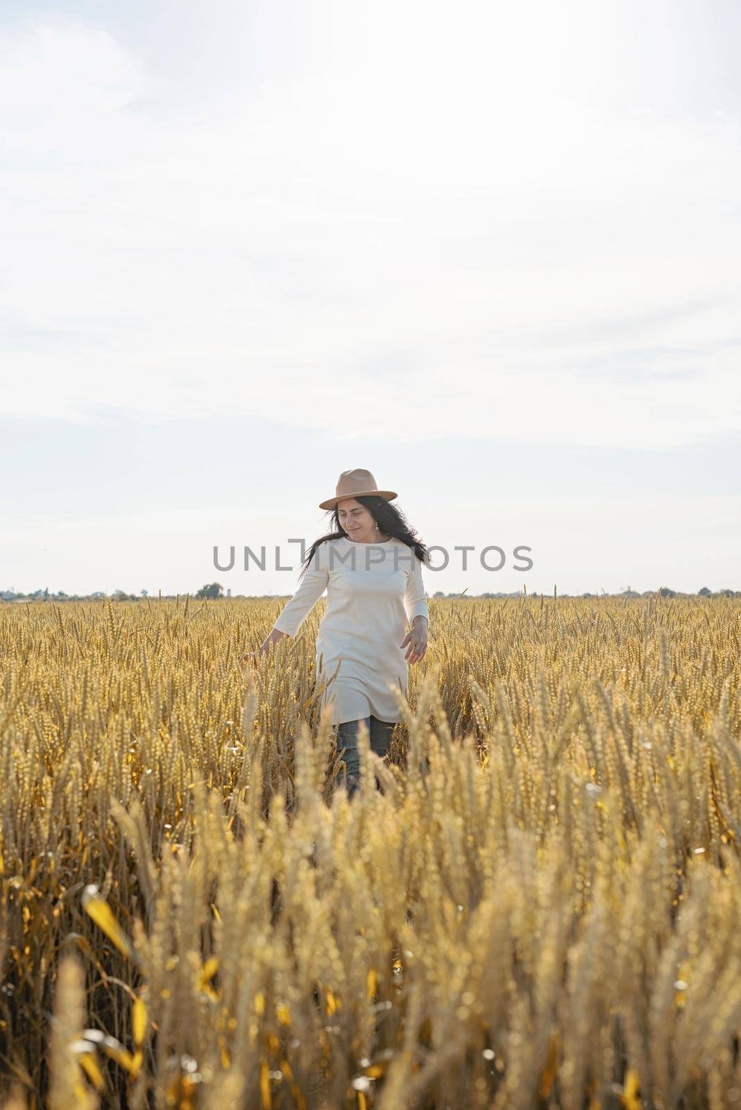 Mid adult woman in white dress walking and dancing across golden field holding heap of rye lit by sunset light, copy space