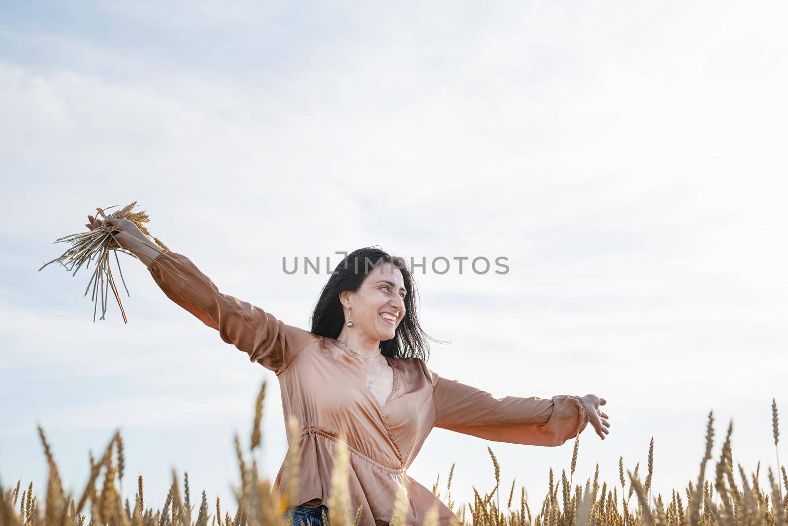 Mid adult woman in beige shirt walking across golden field holding heap of rye lit by sunset light, copy space