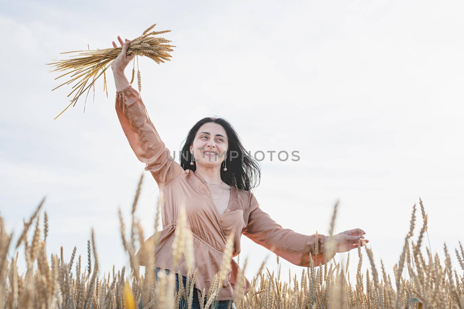 Mid adult woman in beige shirt walking across golden field holding heap of rye lit by sunset light, copy space