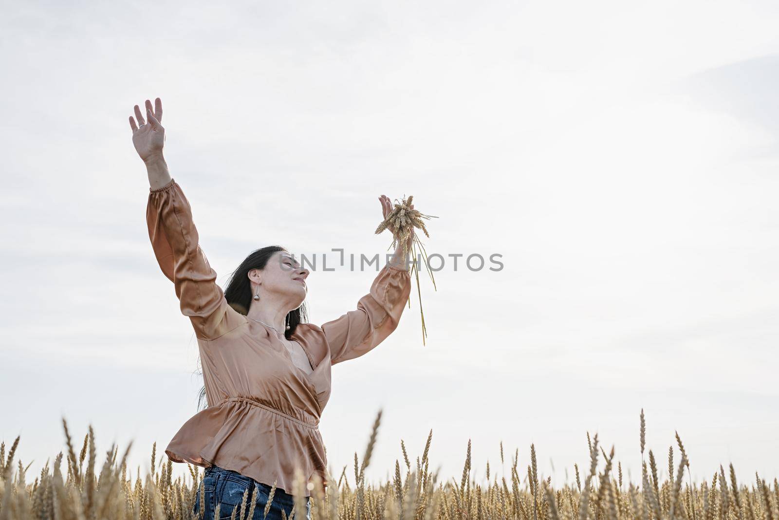 Mid adult woman in beige shirt standing on a wheat field with sunrise on the background, back view by Desperada