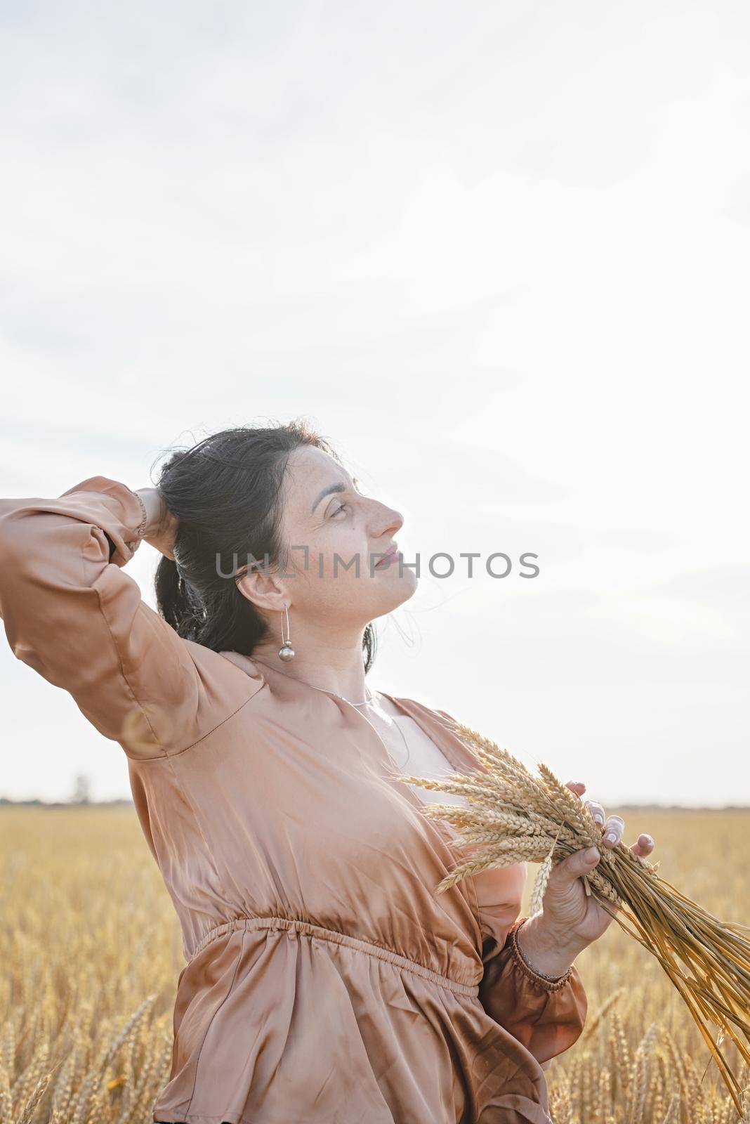 Mid adult woman in beige shirt standing on a wheat field with sunrise on the background, back view by Desperada