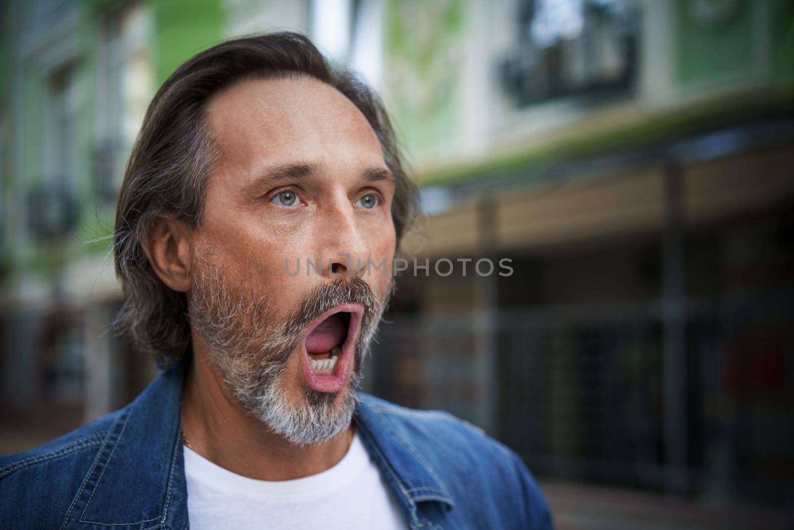 Handsome middle aged european man screaming on the street. Mature male in dressed up in casual jeans shirt stand with open mouth on street of old town. Yarning wide open moth man outdoors.