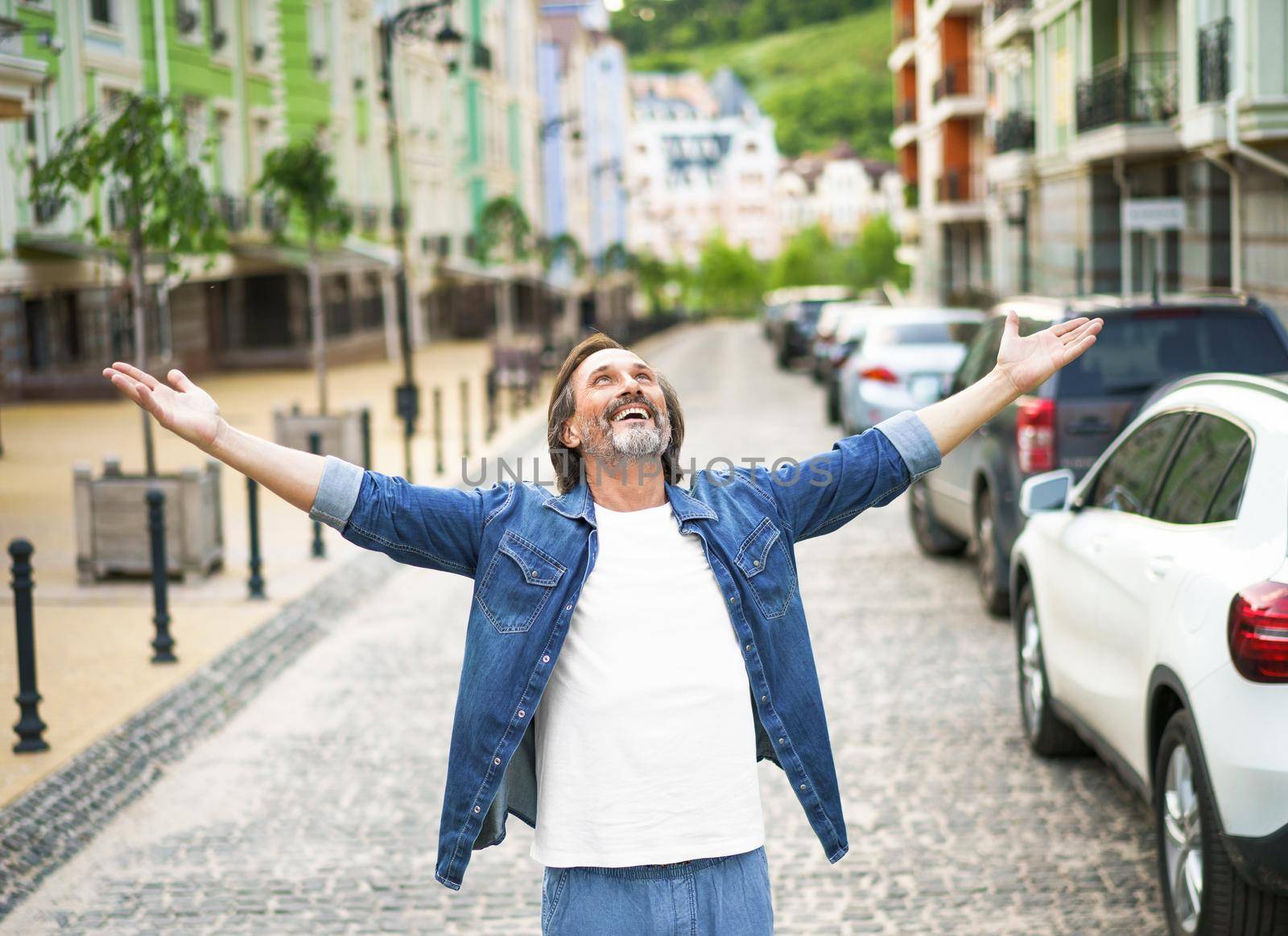 Happy senior man thanks God standing outdoors with hands spread wide open wearing jeans shirt with white t-shirt under. Mid Aged man glad that life is beautiful standing in old town street.