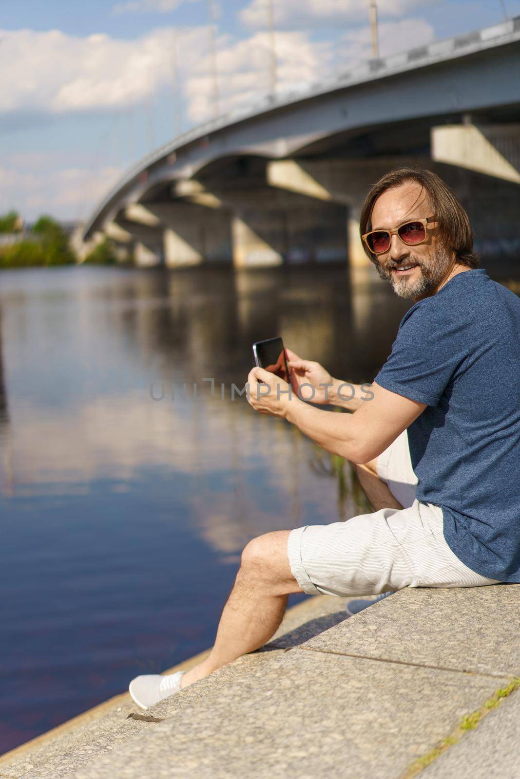 Excited freelancer man sitting outdoors next to river working using digital tablet pc and showing debit or credit card, shopping online or paying online banking. E-banking concept by LipikStockMedia