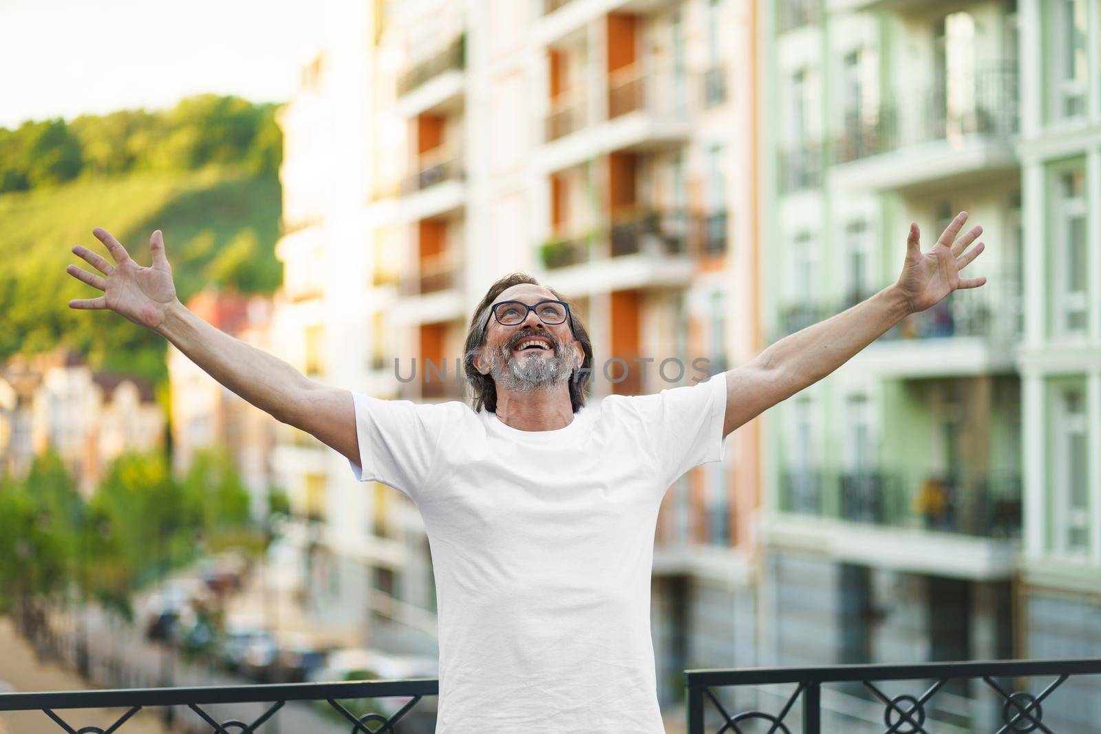 Senior man in glasses praising God standing outdoors with hands spread wide open lifted in the air wearing white t-shirt. Middle aged man glad that life is beautiful standing in old town street.