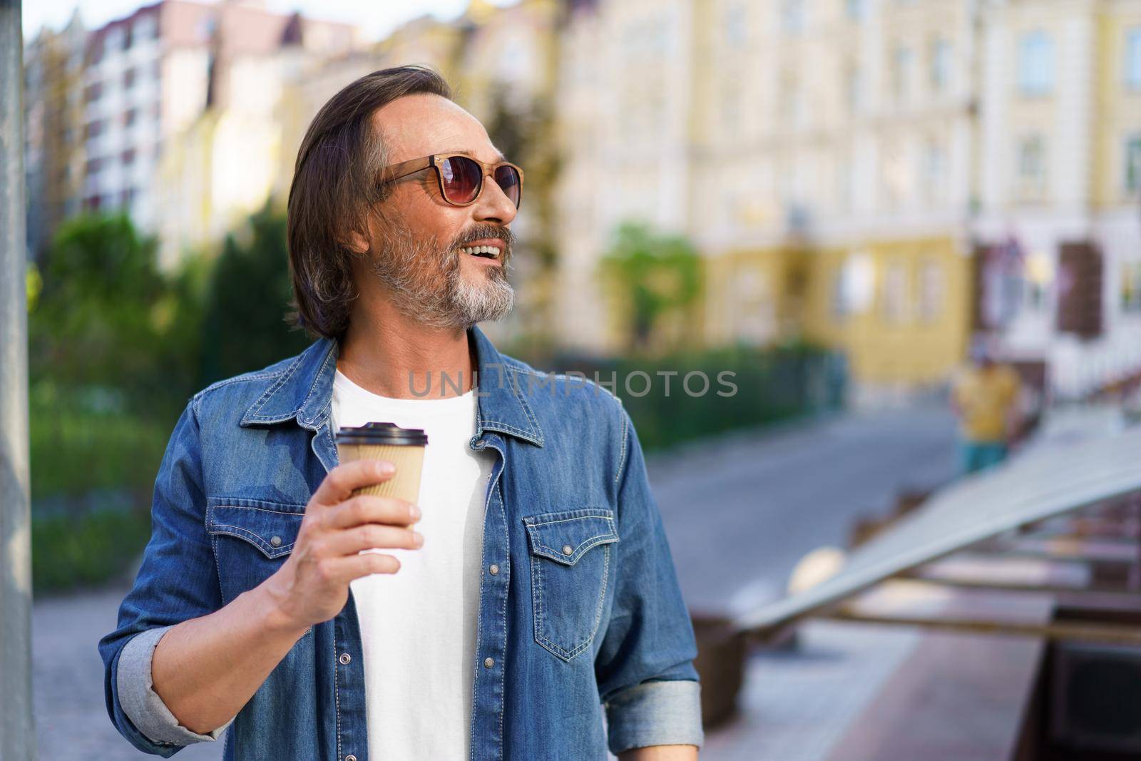 Mature man smiling looking away sideways catching last rays of sun wearing sunglasses with with coffee in paper cup at old town streets. Handsome middle aged man with take away coffee.