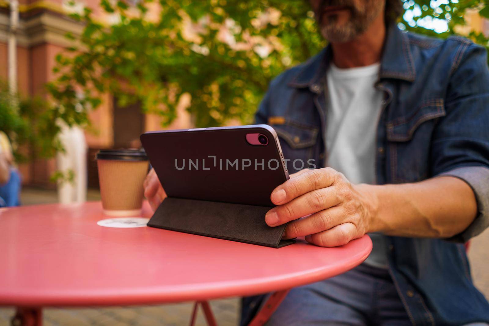 Working outdoors mature bearded man using digital tablet while enjoying coffee. Handsome freelancer traveling man checking on work email or social media sitting at city cafe outdoors. Travel concept.