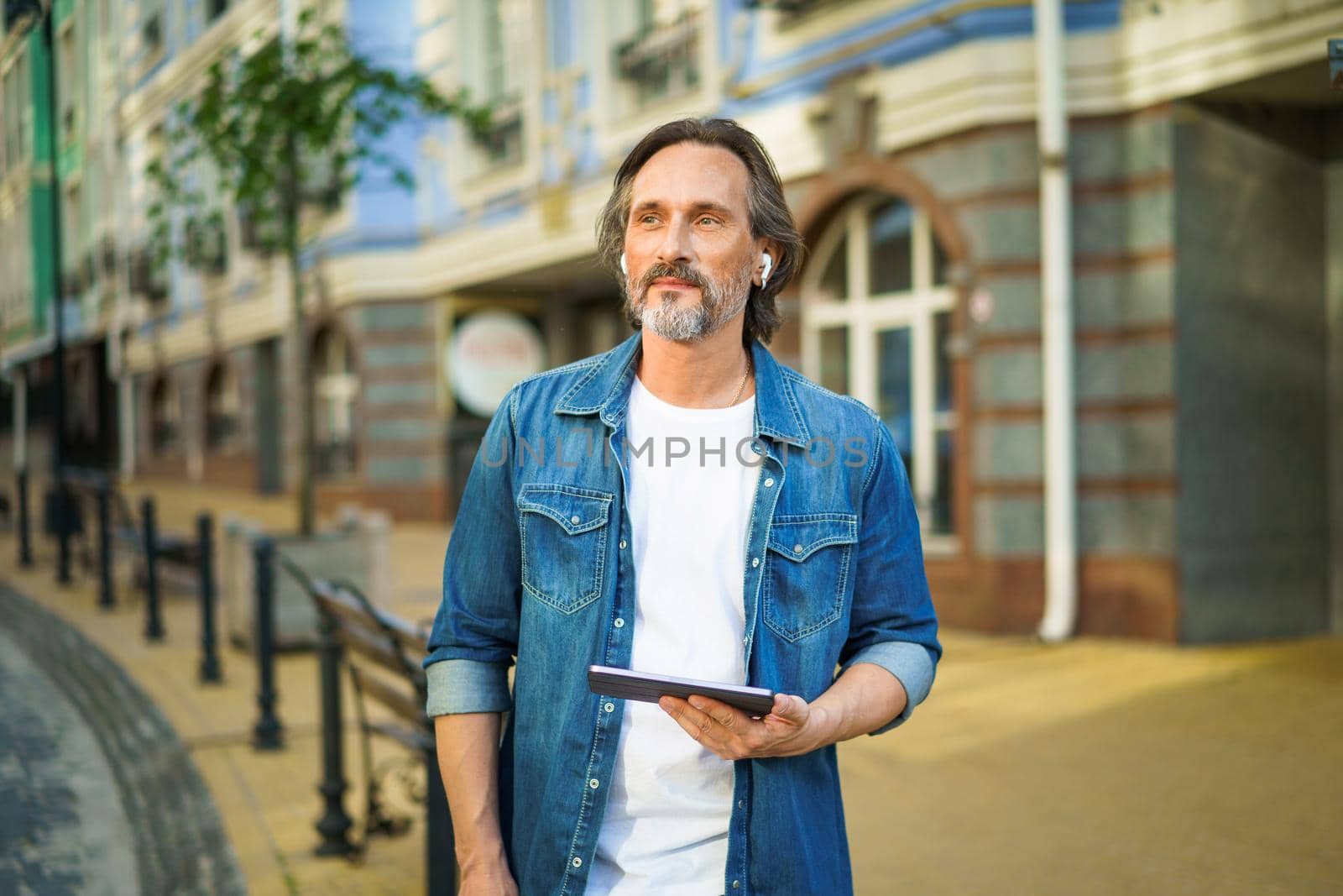 Traveling handsome mature man use digital tablet navigation or having a call while standing on urban streets. Mature man listening music use wireless earphones while travel old town streets by LipikStockMedia