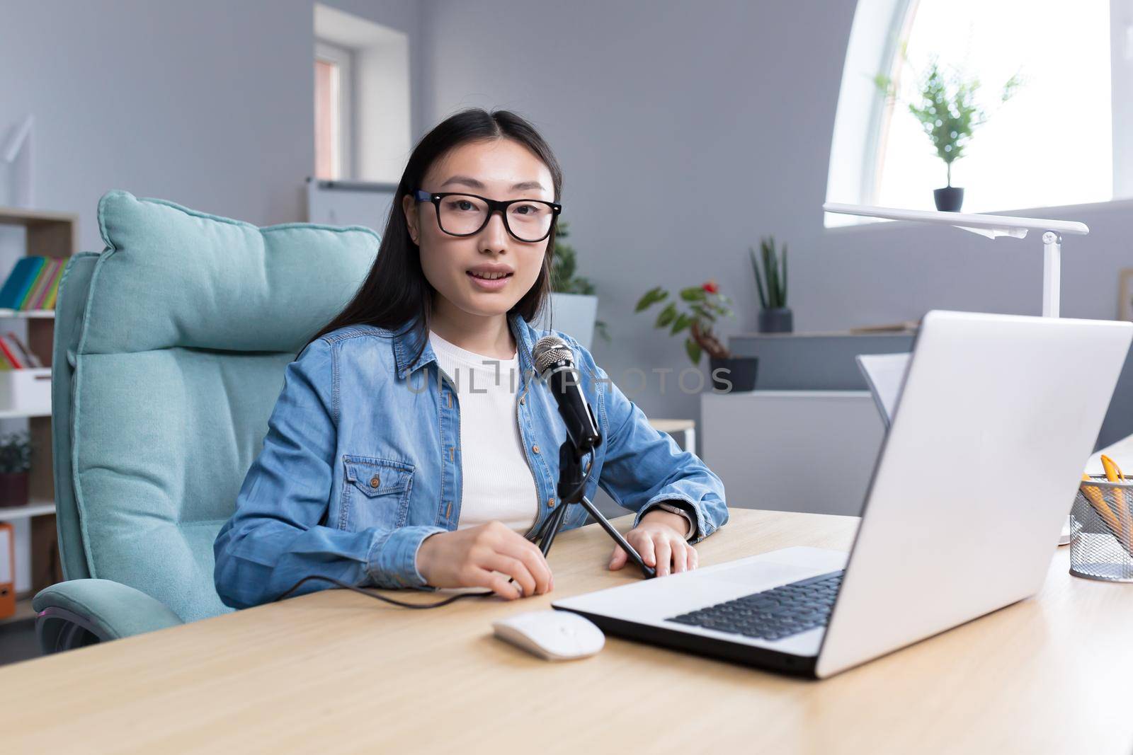 Portrait of young beautiful and successful Asian woman blogger, smiling and looking at camera, working in studio office recording audio podcast, using professional microphone.