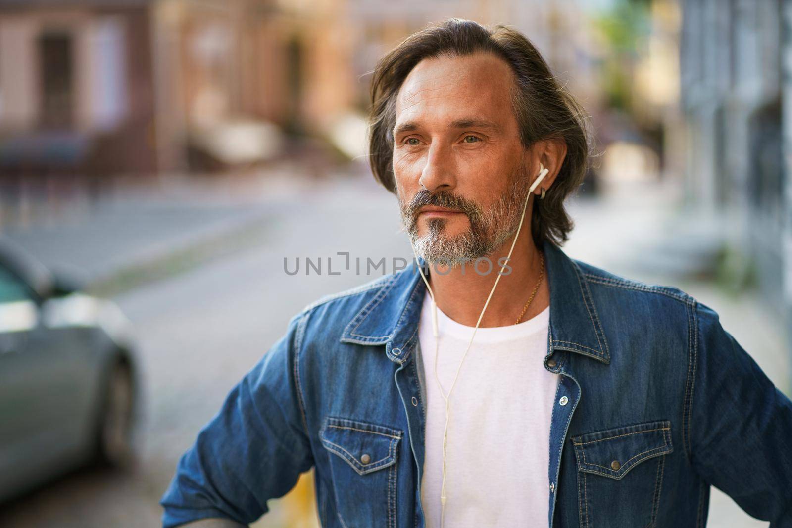 Thoughtful handsome mature grey bearded man with earphones listening music or talking on the phone standing urban street wearing casual jeans shirt with white t-shirt. Travel lifestyle concept.