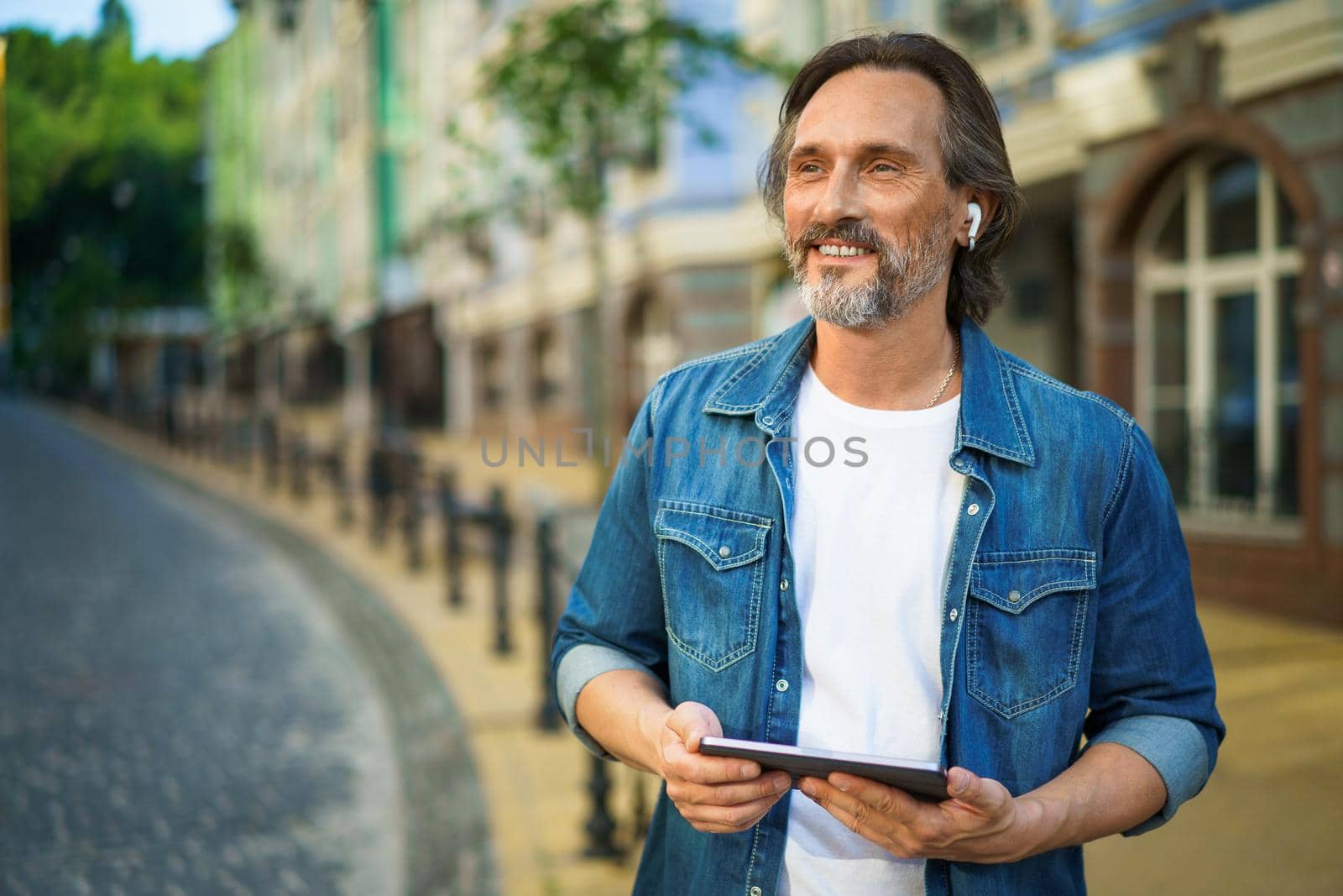 Handsome mature freelancer man with digital tablet in hands having a call while standing on urban streets. Mature man talking or listening music use wireless earphones while travel old town streets.