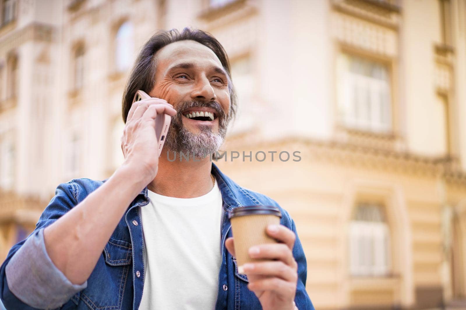 Happy traveling mature bearded man talking using mobile phone and drinking coffee using disposable paper cup standing outdoors in old city background wearing jeans shirt. Freelancer traveling man.