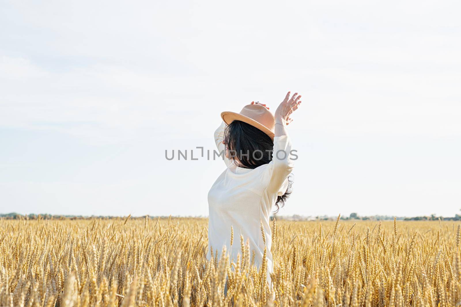 Mid adult woman in white dress walking and dancing across golden field holding heap of rye lit by sunset light, copy space