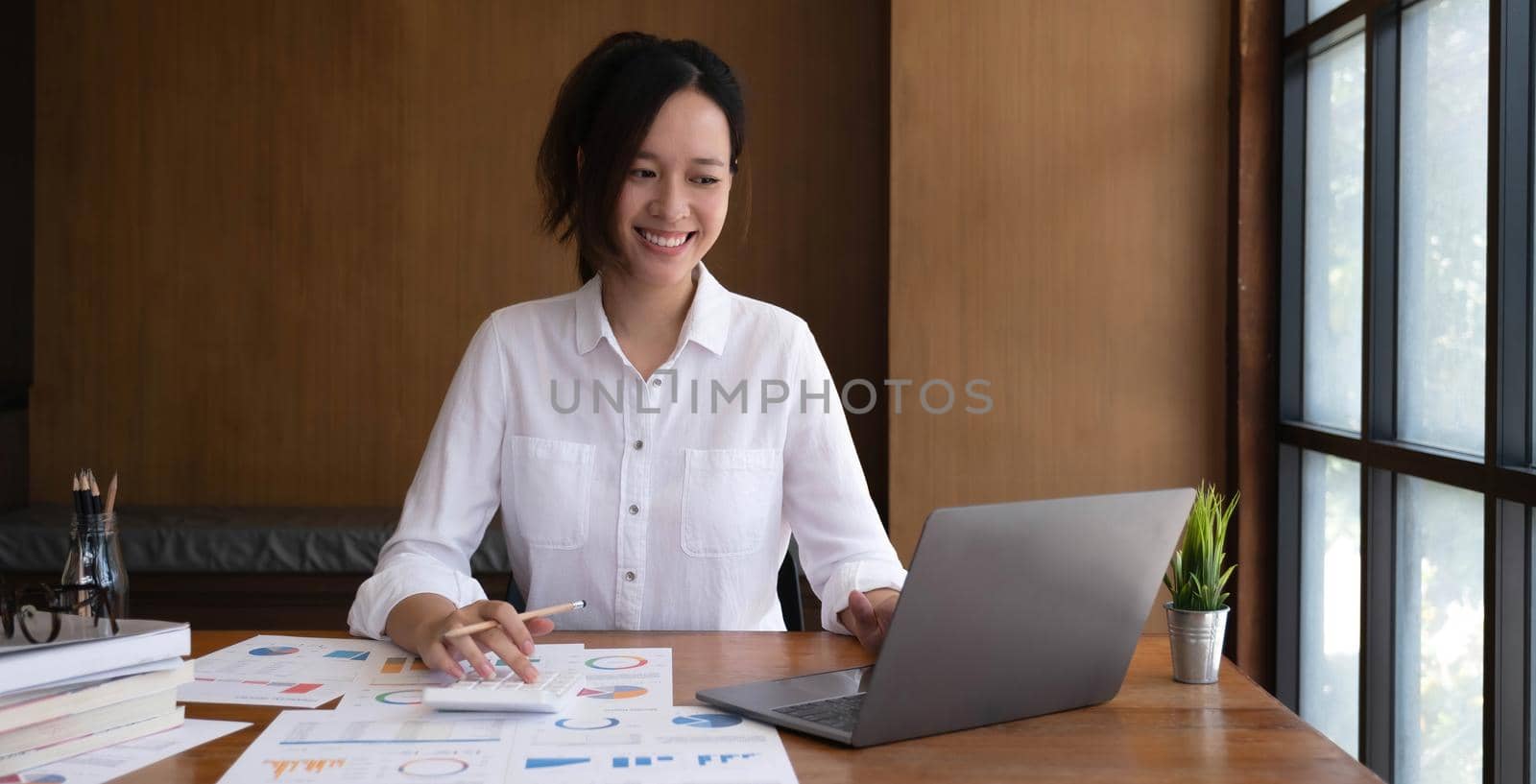 Smiling beautiful young asian woman working on laptop computer while sitting at the cafe indoors, looking through documents by wichayada