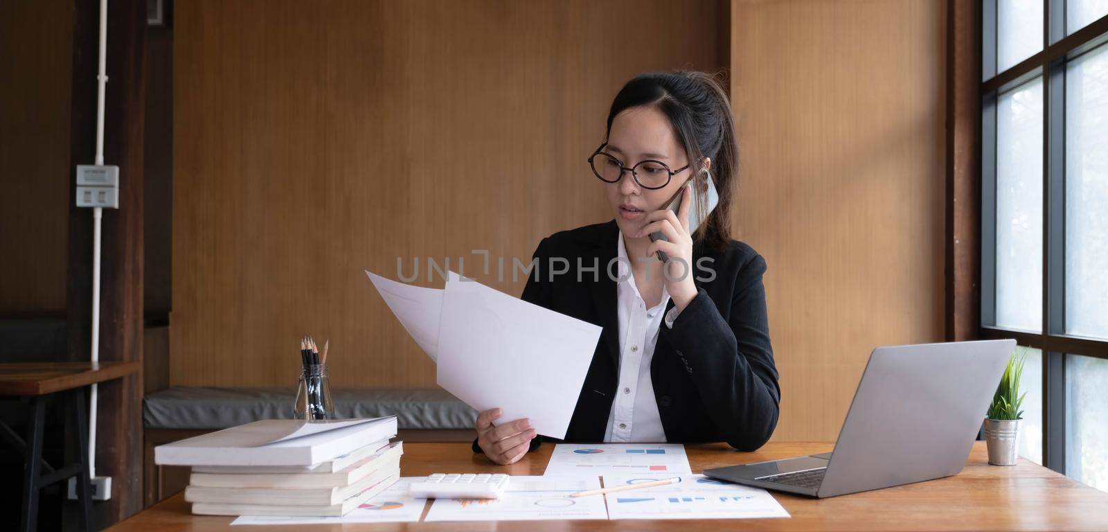 Serious asian businesswoman using laptop looking at computer talking on phone consult client sit at desk, focused female sales manager make mobile business call discuss work with customer in office by wichayada