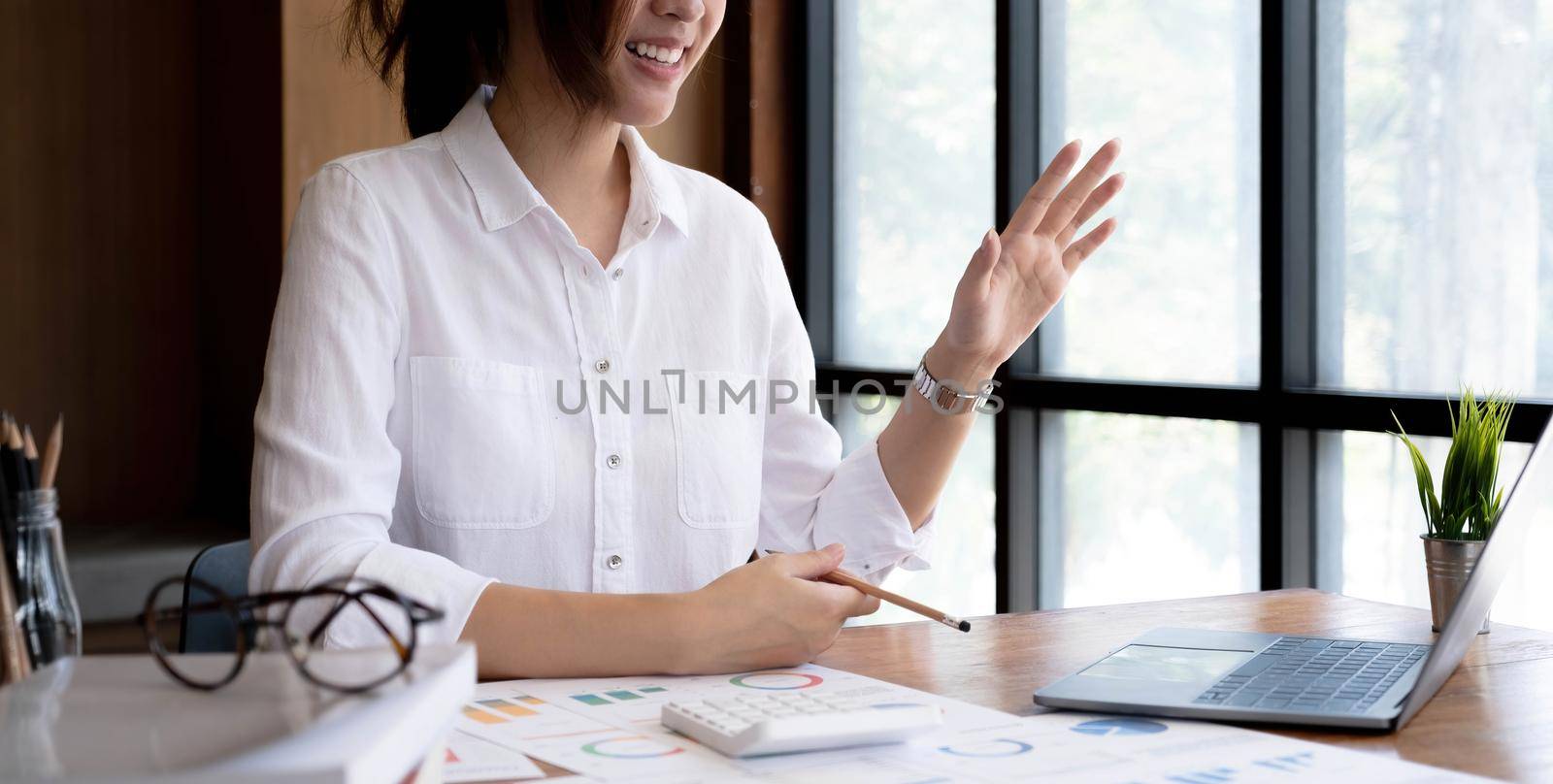 happy and smiling young asian corporate executive working from home greeting colleagues or clients during meeting using video call.