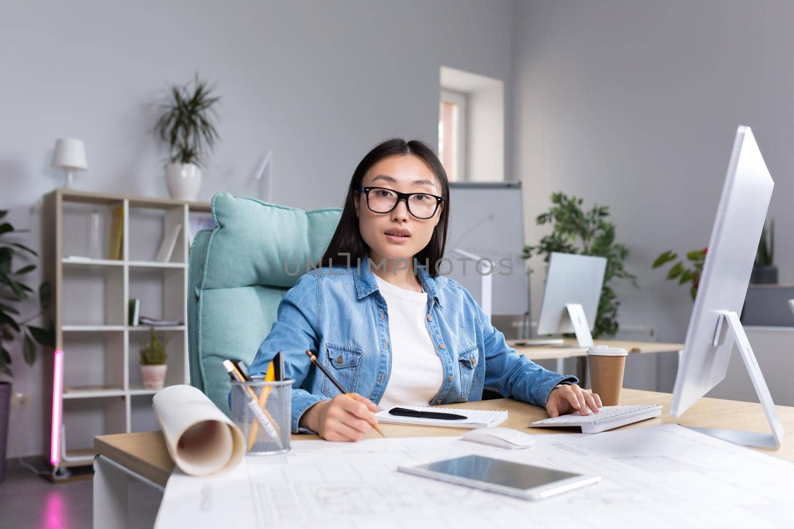 Portrait of young designer woman in modern office at work, successful Asian woman in glasses looking at camera.