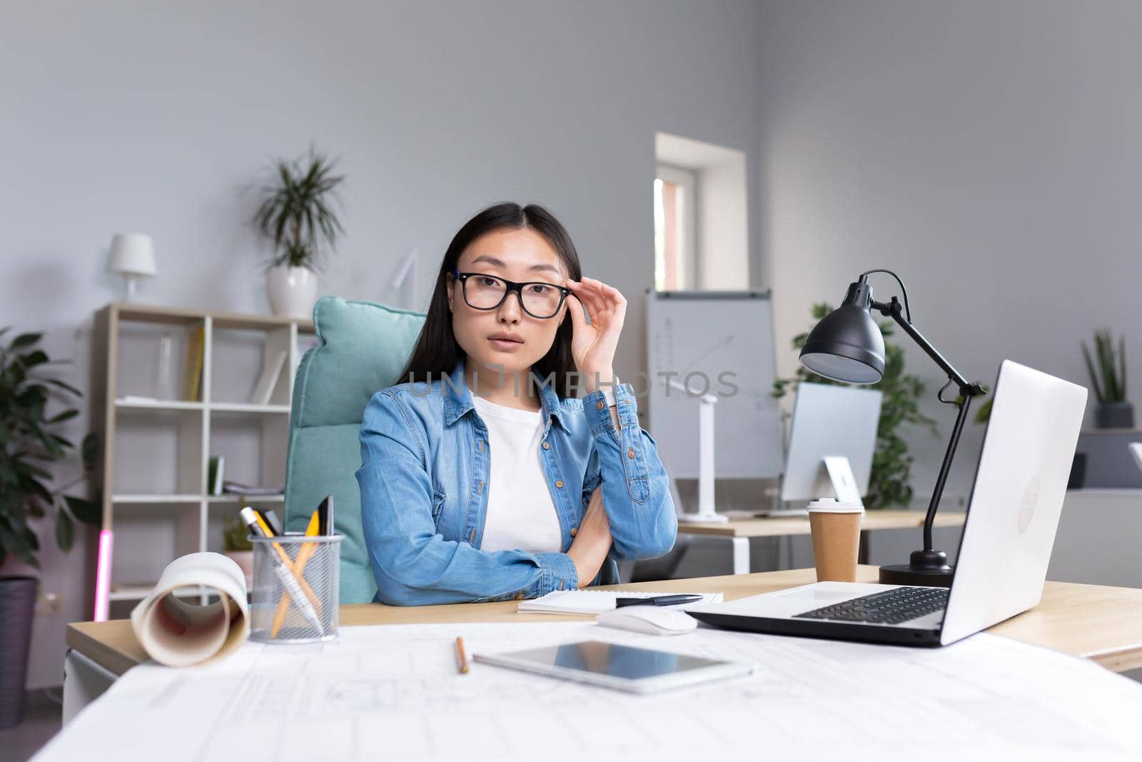 Portrait of young designer woman in modern office at work, successful Asian woman in glasses looking at camera.