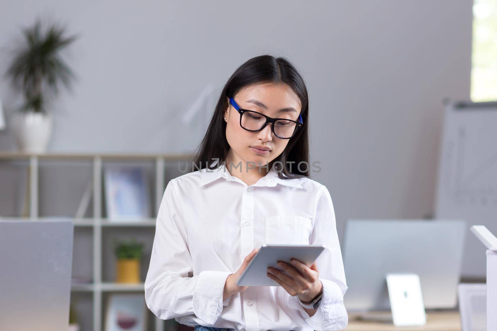 Young beautiful Asian teacher working with tablet, in classroom with computers at school, woman in white shirt and glasses reads from tablet