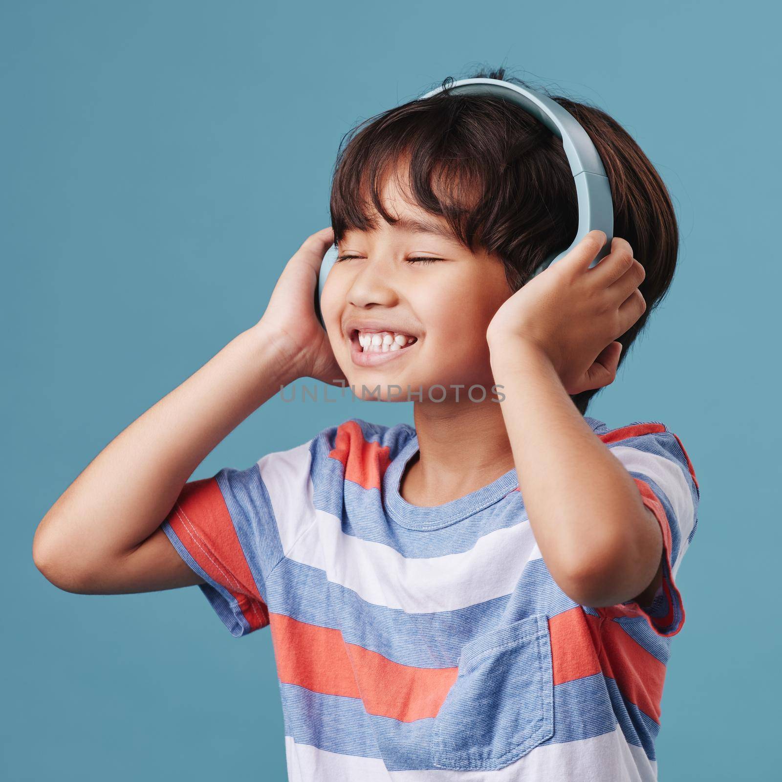 A cute young asian boy enjoying listening to music from his headphones. Adorable Chinese kid feeling the magic of music while posing against a blue studio background.