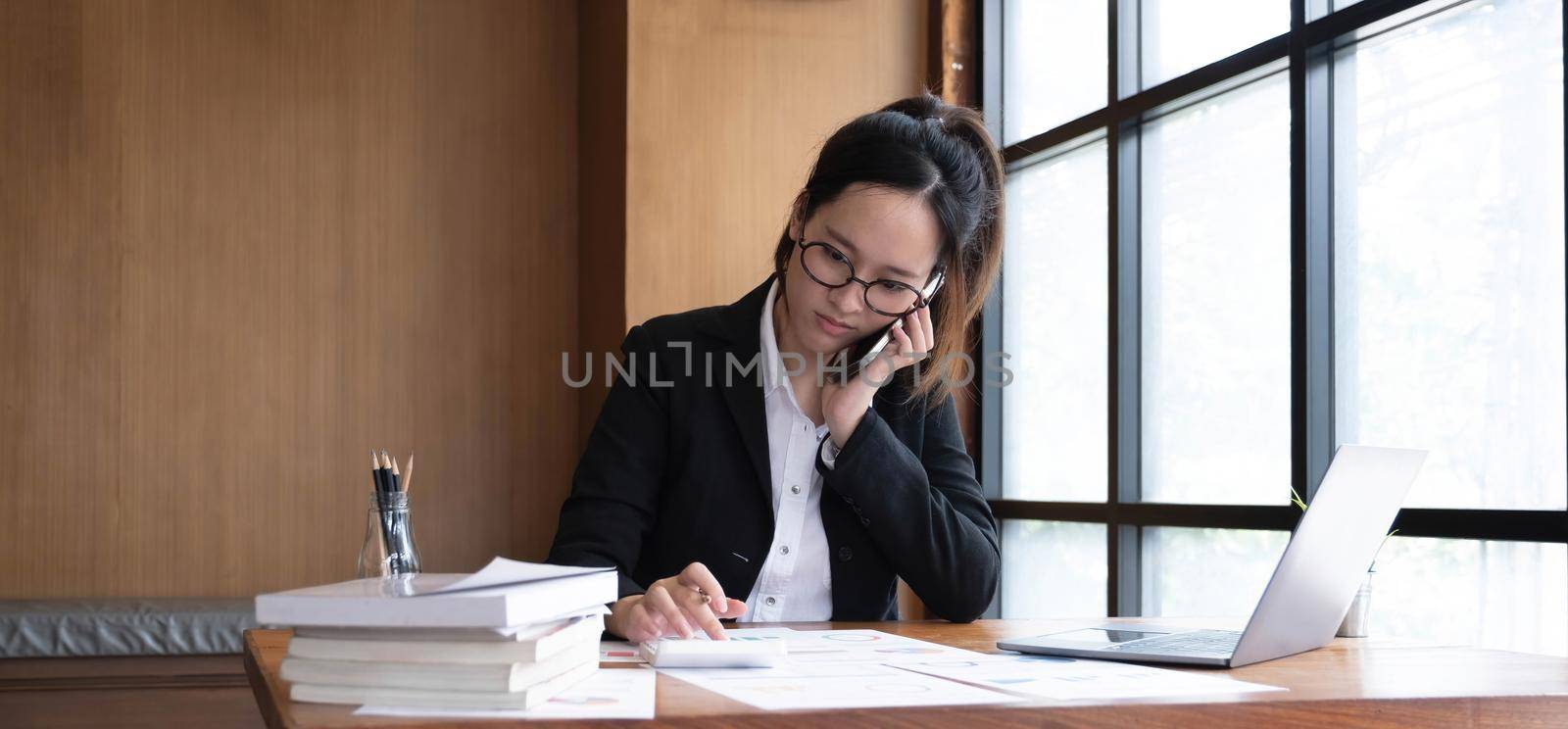 Serious asian businesswoman using laptop looking at computer talking on phone consult client sit at desk, focused female sales manager make mobile business call discuss work with customer in office by wichayada