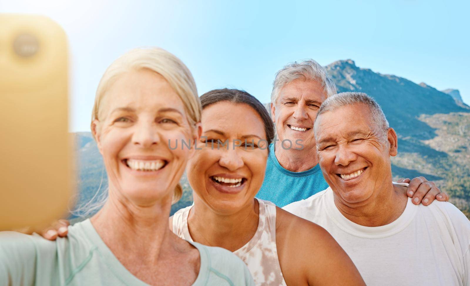 Group of active seniors posing together for a selfie or video call on a sunny day against a mountain view background. Happy retirees exercising together outdoors. Living healthy and active lifestyles by YuriArcurs