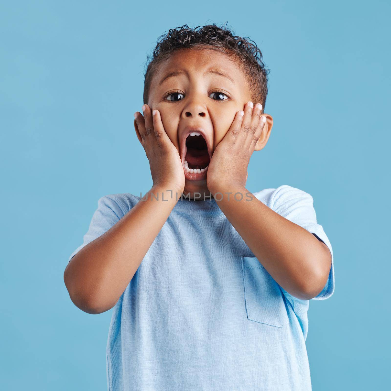 Adorable little hispanic boy with hands on face and mouth open looking terrified and shocked showing true astonished reaction against a blue studio background.