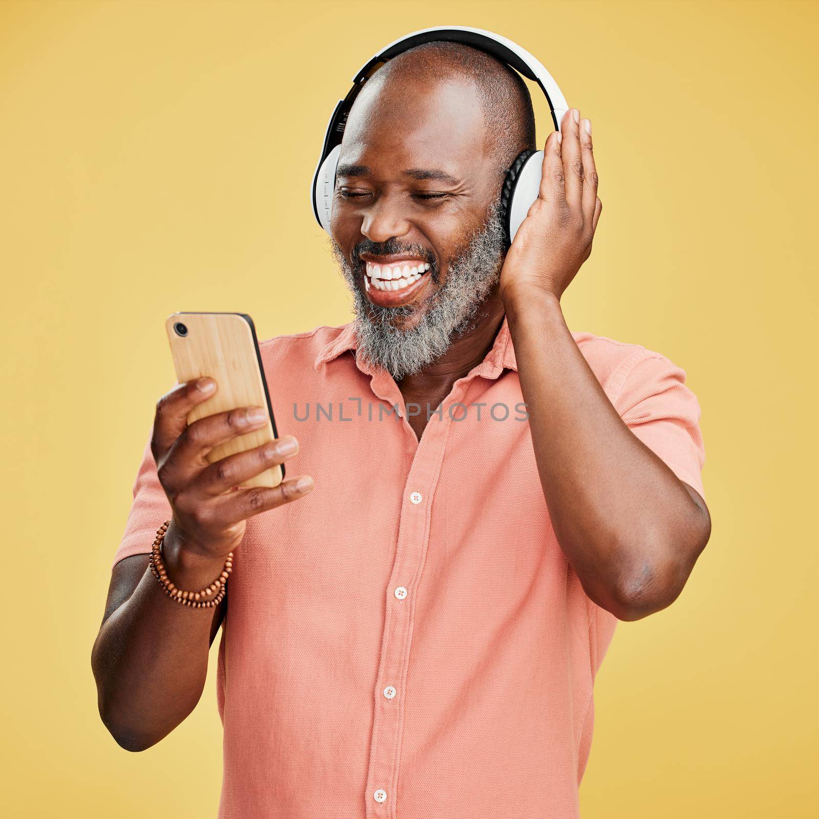 One mature african american man listening to music using wireless headphones while isolated against a yellow background. Happy man with a grey beard smiling while streaming on his phone in studio by YuriArcurs