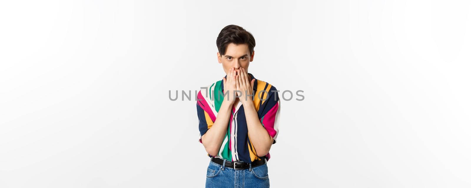 Image of young gay man looking intrigued, holding hands on mouth, gossiping, standing over white background by Benzoix