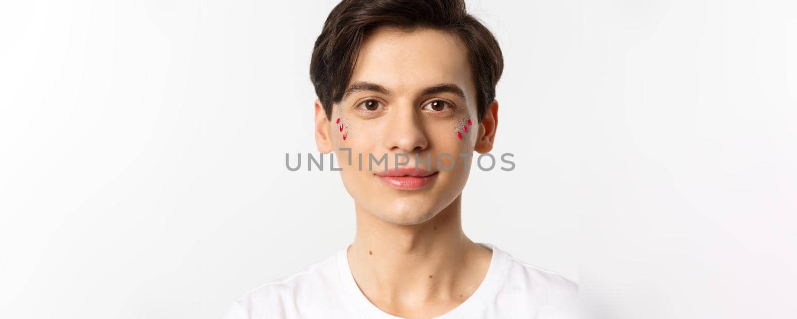 People, lgbtq and beauty concept. Close-up of happy queer guy with applied lip gloss and glitter, smiling and looking at camera, standing over white background.