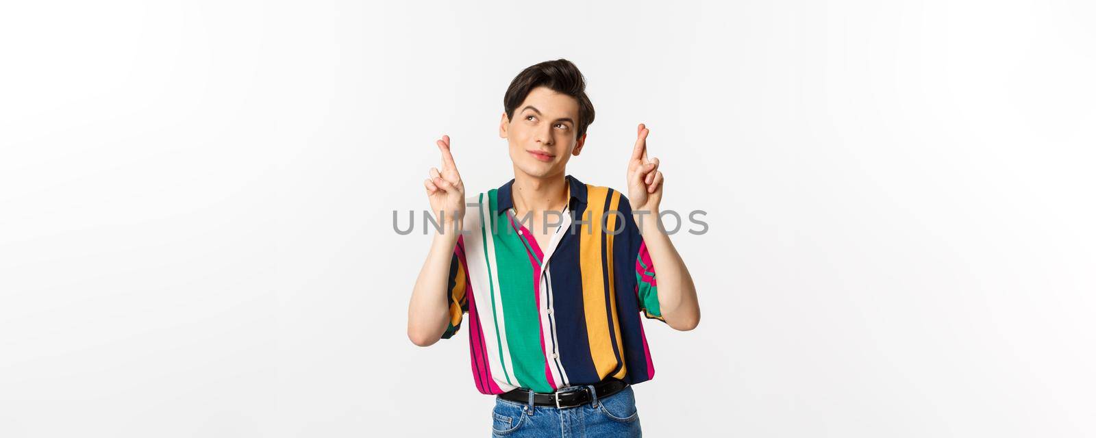 Dreamy young man making wish with fingers crossed, smiling and looking at upper left corner thoughtful, standing over white background by Benzoix