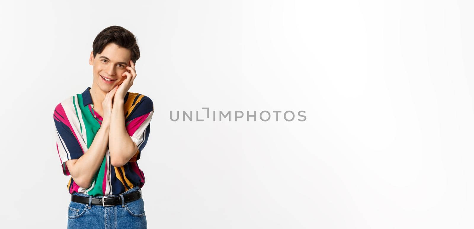 Beautiful young man looking with admiration at camera, smiling, gently touching face, standing over white background.