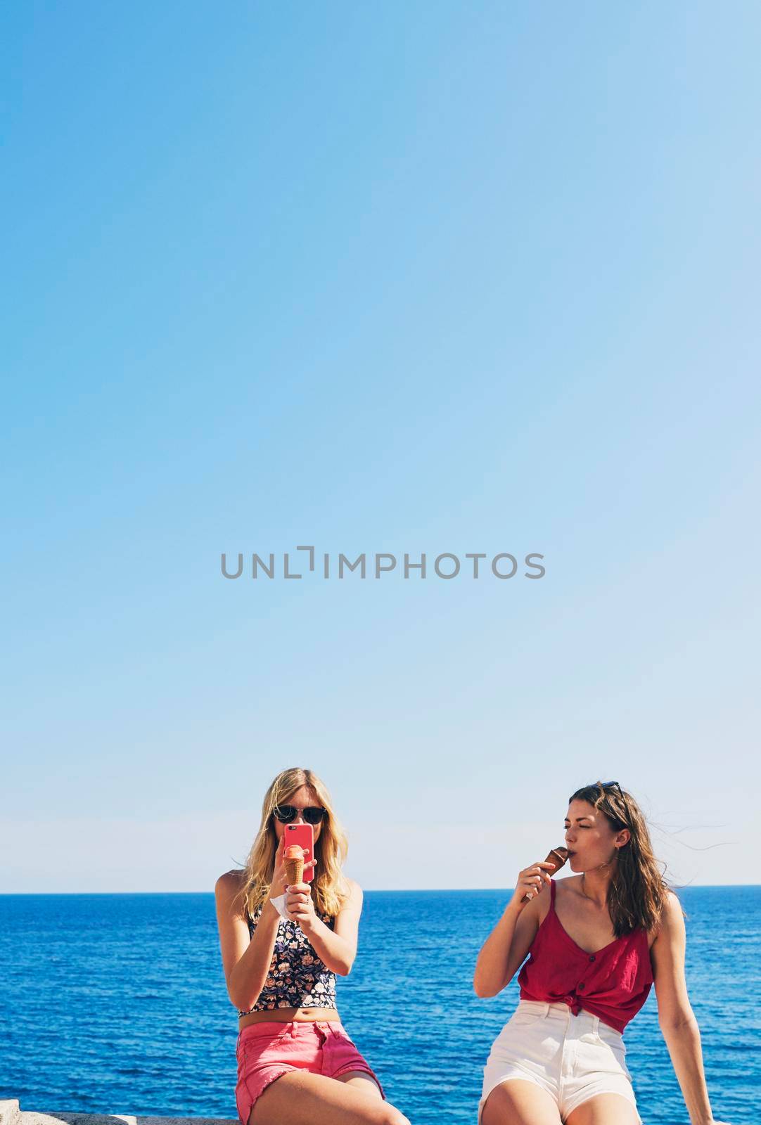 Two friends taking photos of ice cream eating on beach using smart phone technology on adventure travel by YuriArcurs