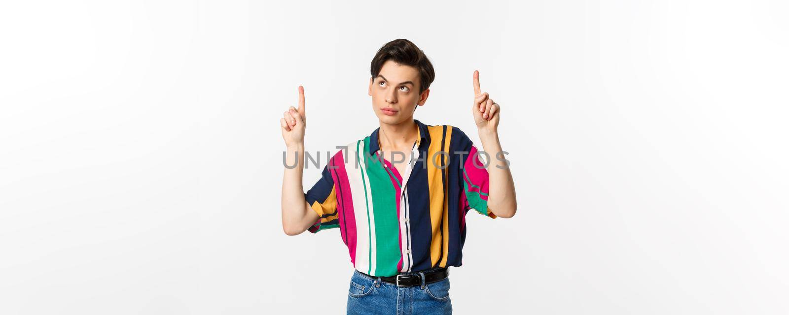 Attractive gay man looking thoughtful, pointing fingers up at logo, standing over white background. Copy space